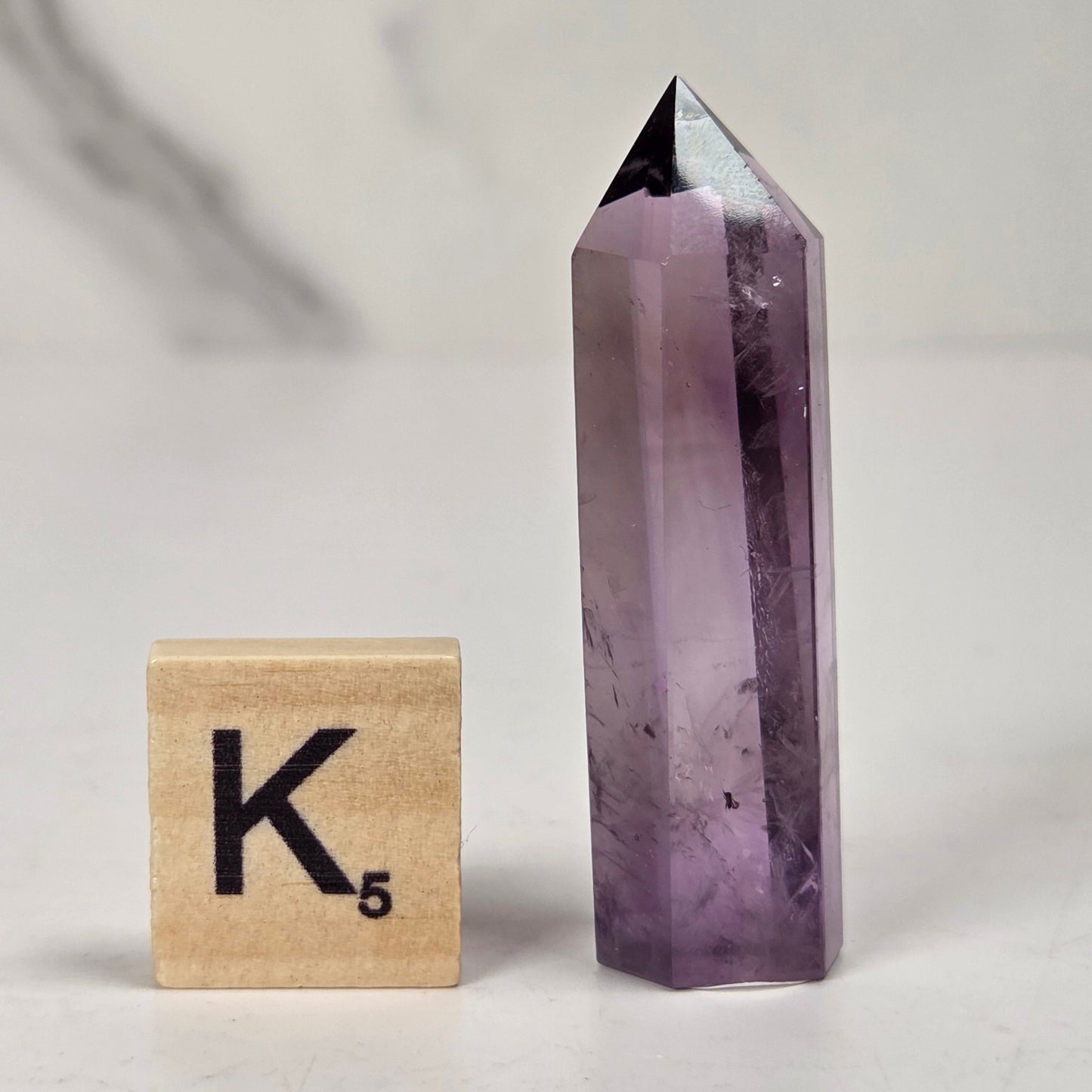 A pointed amethyst crystal tower stands next to a wooden Scrabble tile showing the letter K and value 5, set against a light gray marble-patterned background.