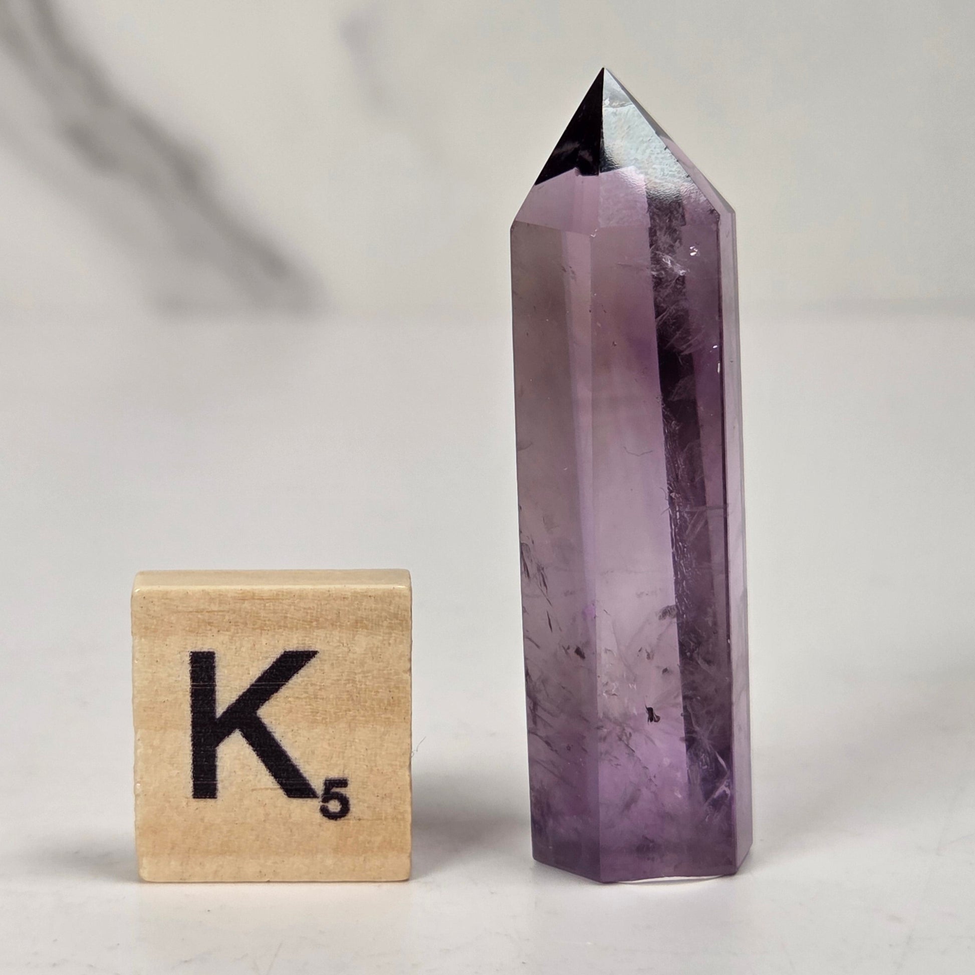 A pointed amethyst crystal tower stands next to a wooden Scrabble tile showing the letter K and value 5, set against a light gray marble-patterned background.