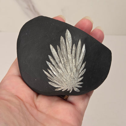 A hand holding a black slate stone featuring a textured, metallic silver fern-like design in the center, set against a plain light background.