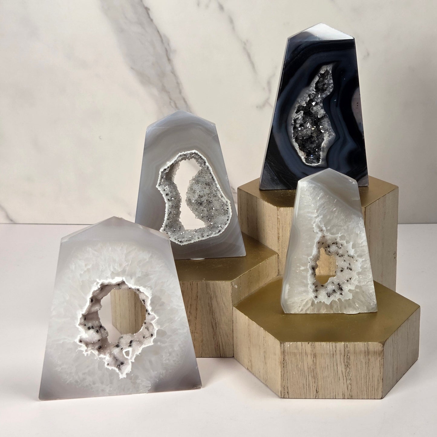Four polished agate geode slices with natural crystal cavities, displayed on wooden hexagonal and rectangular stands against a light marble-patterned background.