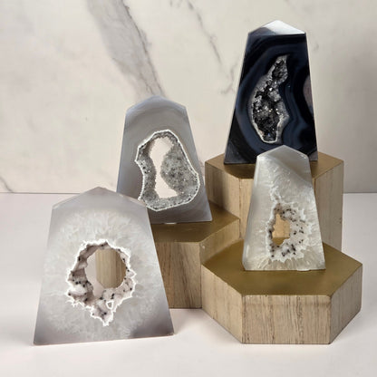Four polished agate geode slices with natural crystal cavities, displayed on wooden hexagonal and rectangular stands against a light marble-patterned background.