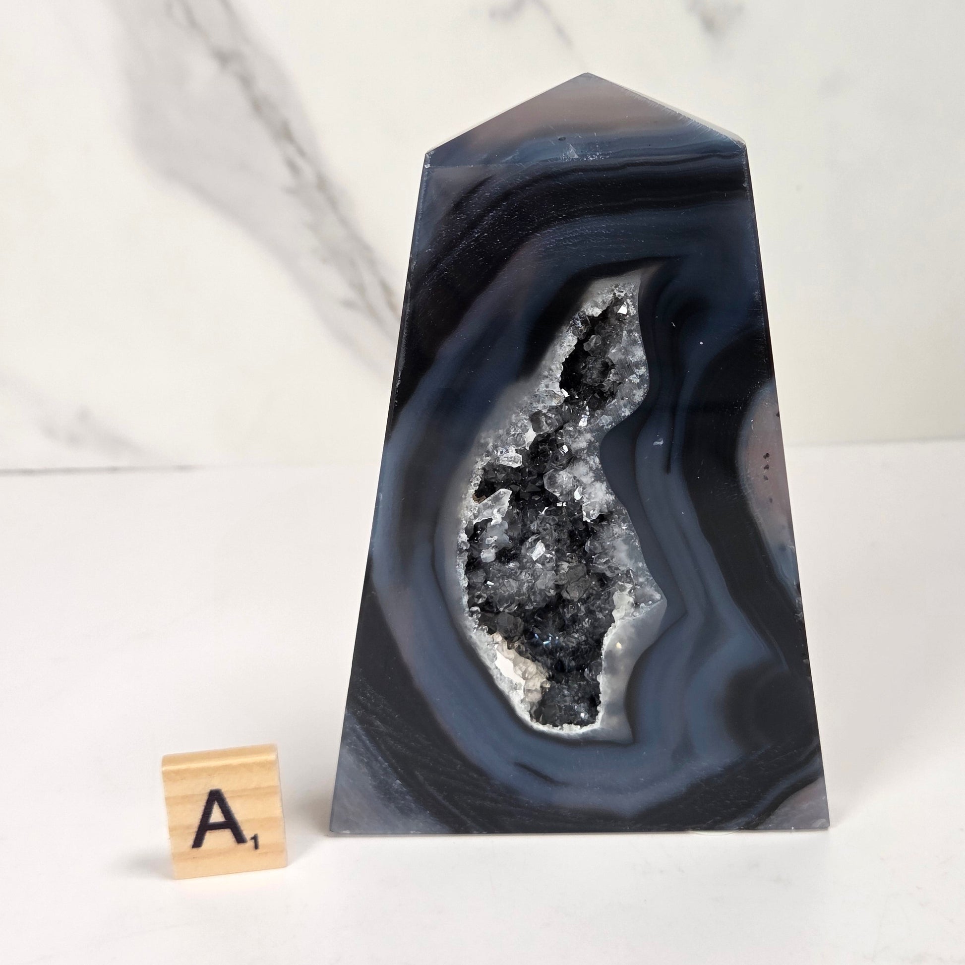 A polished agate crystal point with a black and gray banded outer layer and a white crystalline geode interior, displayed on a white marble surface with a small wooden letter tile.