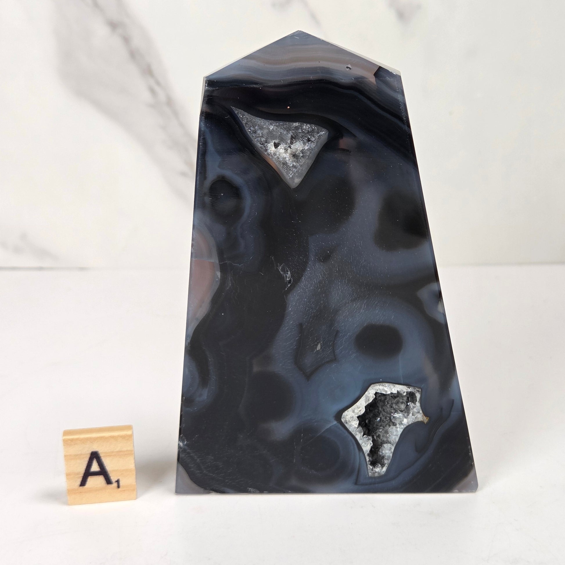 A polished agate crystal point with a black and gray banded outer layer and a white crystalline geode interior, displayed on a white marble surface with a small wooden letter tile.