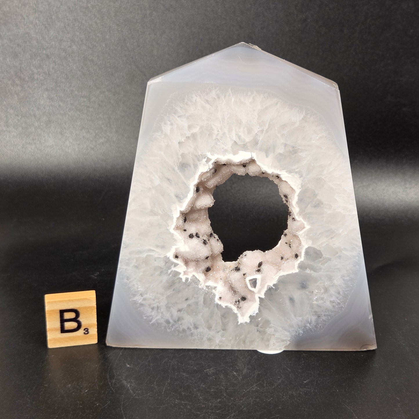 A polished agate crystal point with a black and gray banded outer layer and a white crystalline geode interior, displayed on a black surface with a small wooden letter tile.