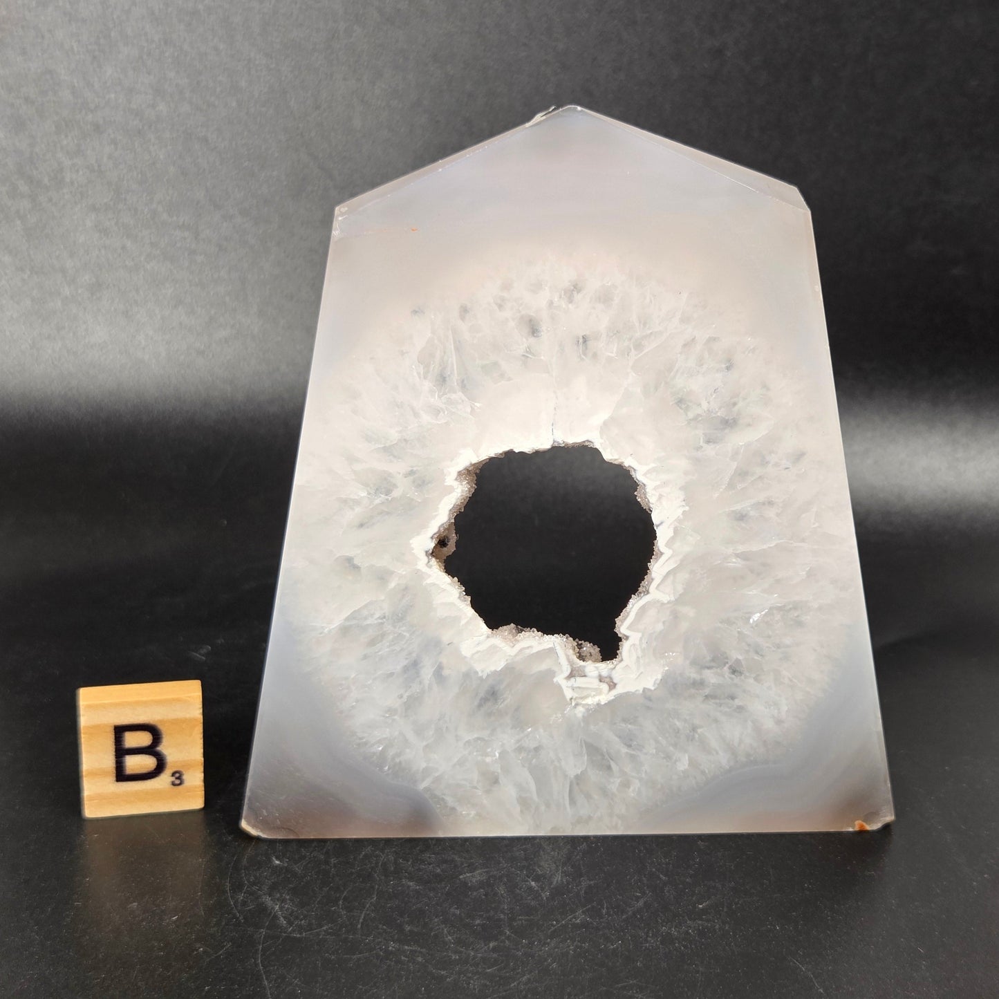 A polished agate crystal point with a black and gray banded outer layer and a white crystalline geode interior, displayed on a black surface with a small wooden letter tile.