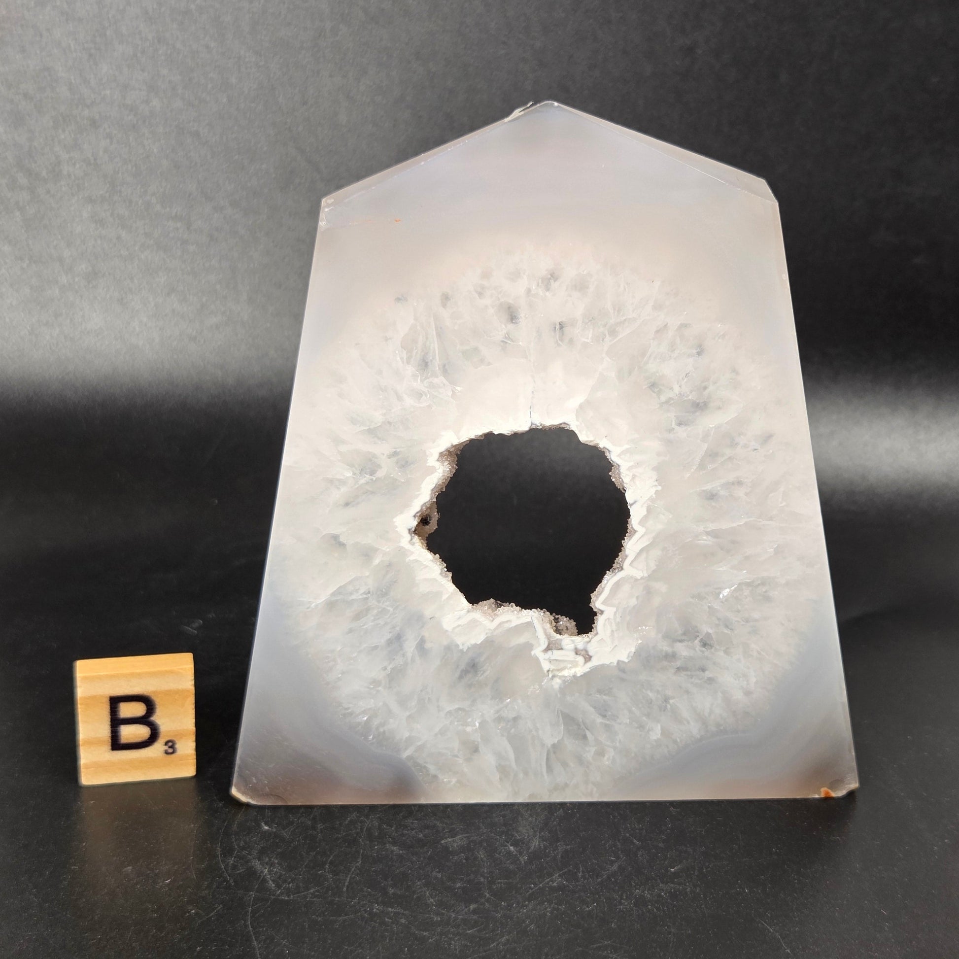 A polished agate crystal point with a black and gray banded outer layer and a white crystalline geode interior, displayed on a black surface with a small wooden letter tile.