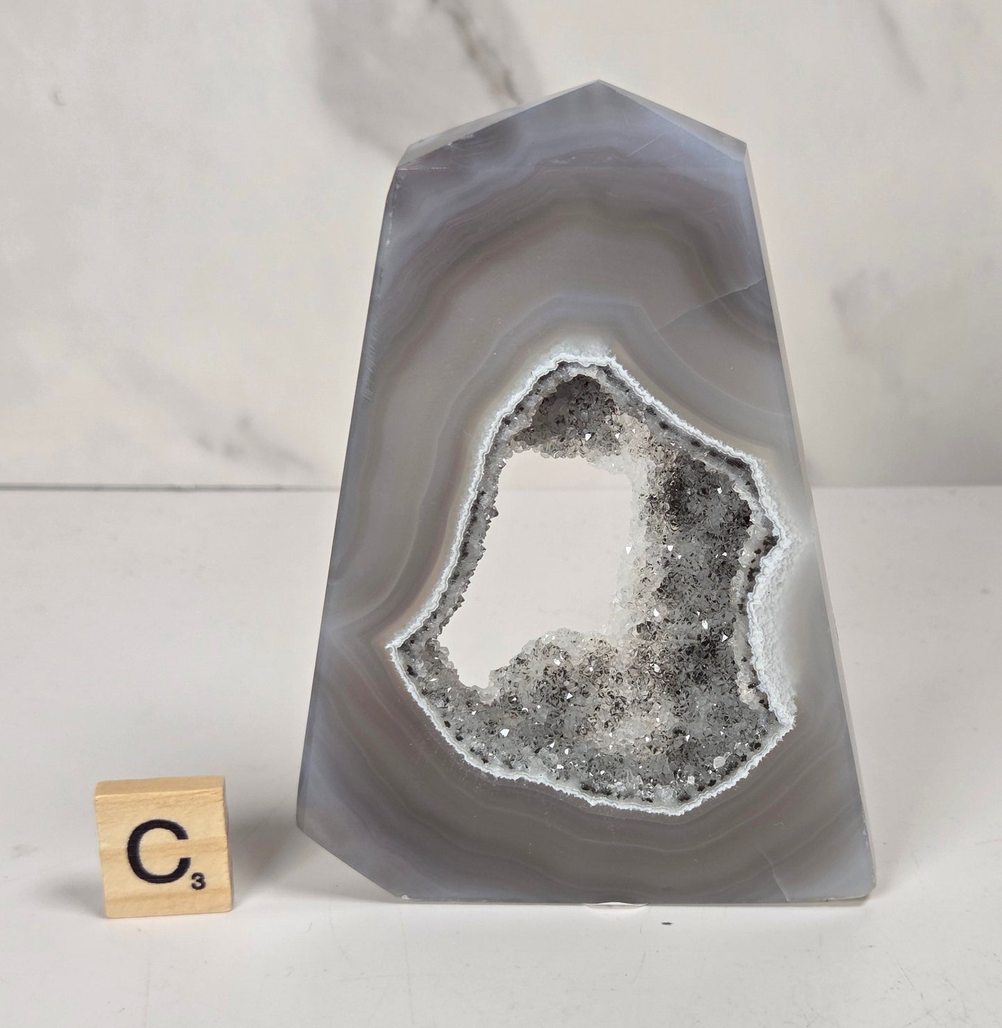 A polished agate crystal point with a black and gray banded outer layer and a white crystalline geode interior, displayed on a marbled surface with a small wooden letter tile.