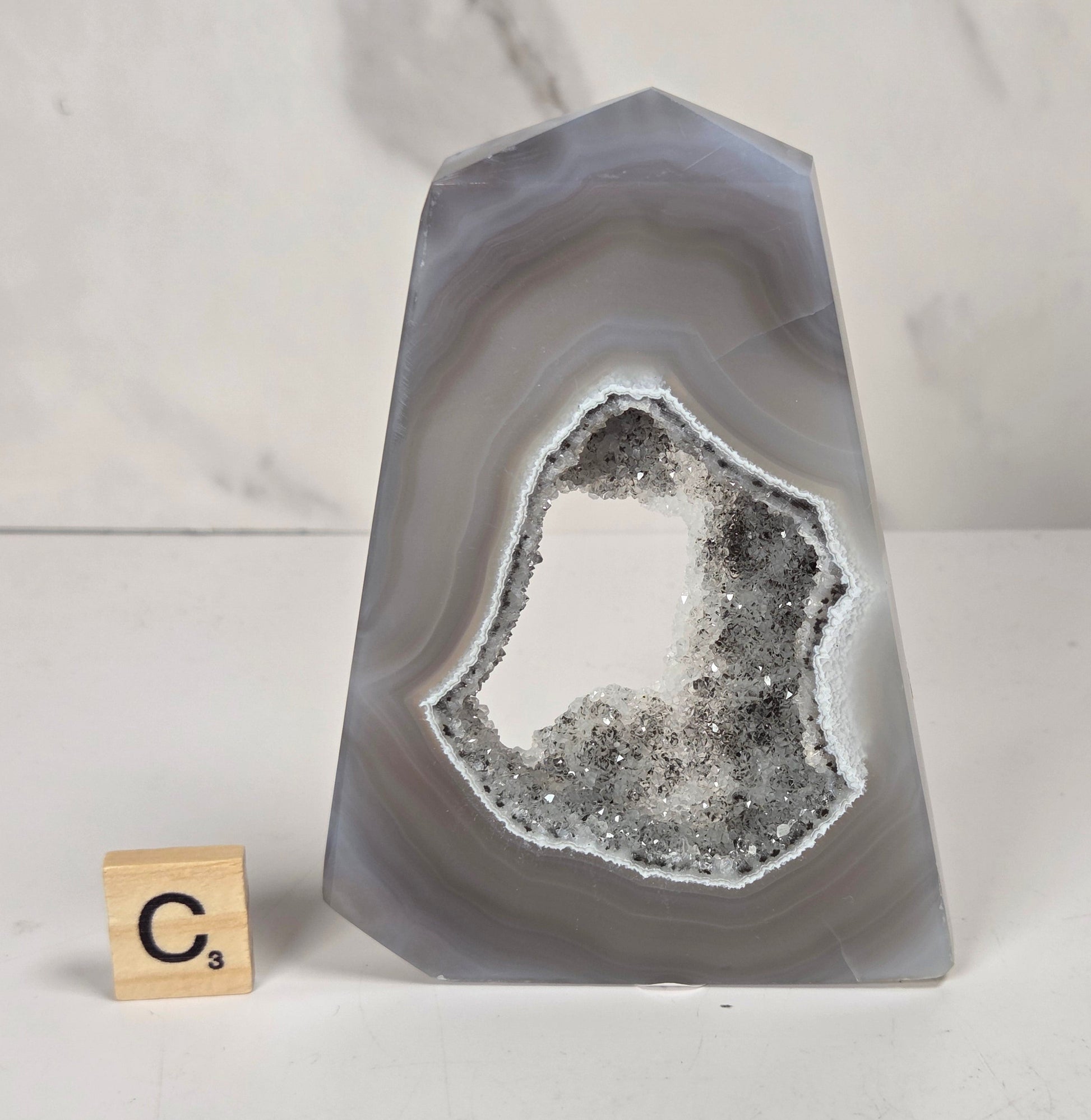A polished agate crystal point with a black and gray banded outer layer and a white crystalline geode interior, displayed on a marbled surface with a small wooden letter tile.