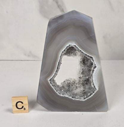 A polished agate crystal point with a black and gray banded outer layer and a white crystalline geode interior, displayed on a marbled surface with a small wooden letter tile.