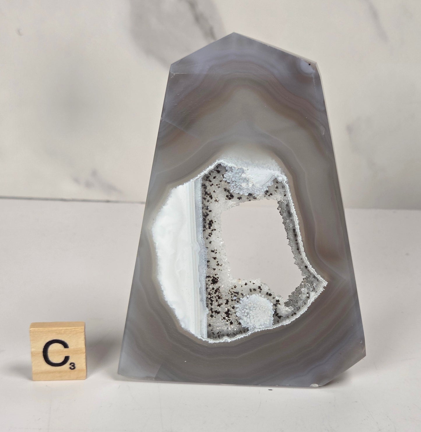 A polished agate crystal point with a natural hollow interior showing white and gray geode formations, displayed on a white surface next to a small Scrabble tile showing the letter C.