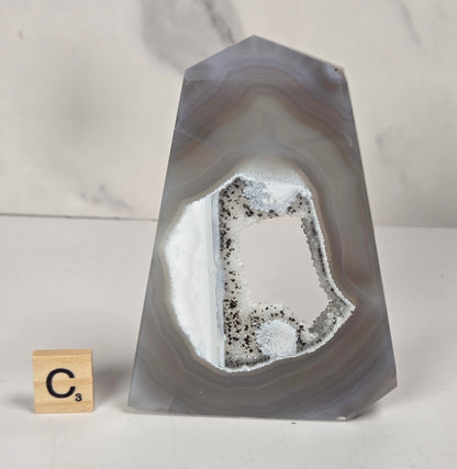 A polished agate crystal point with a natural hollow interior showing white and gray geode formations, displayed on a white surface next to a small Scrabble tile showing the letter C.