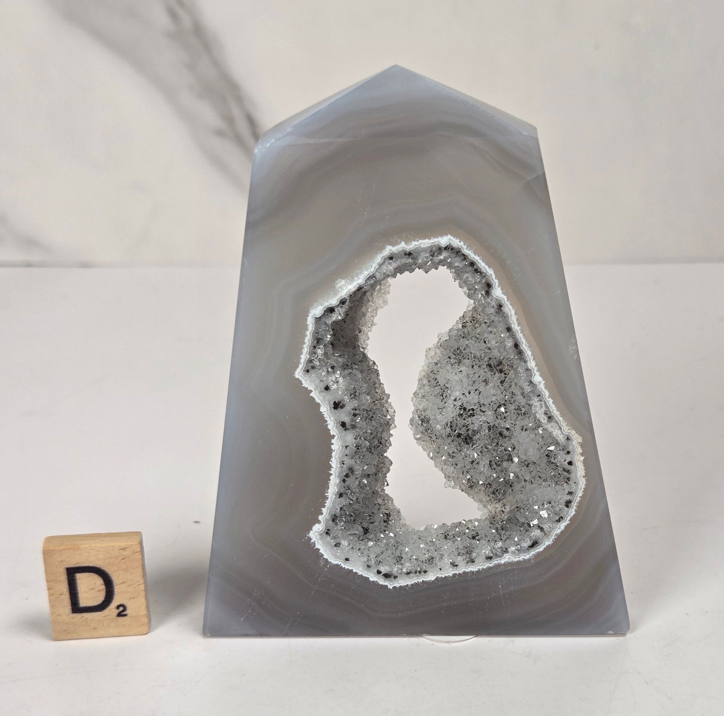 A polished agate crystal tower with a natural geode cavity filled with white and gray crystalline structure, shown on a white surface with a small Scrabble tile for scale.