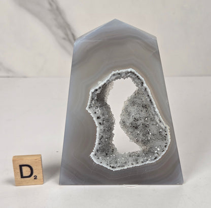 A polished agate crystal tower with a natural geode cavity filled with white and gray crystalline structure, shown on a white surface with a small Scrabble tile for scale.