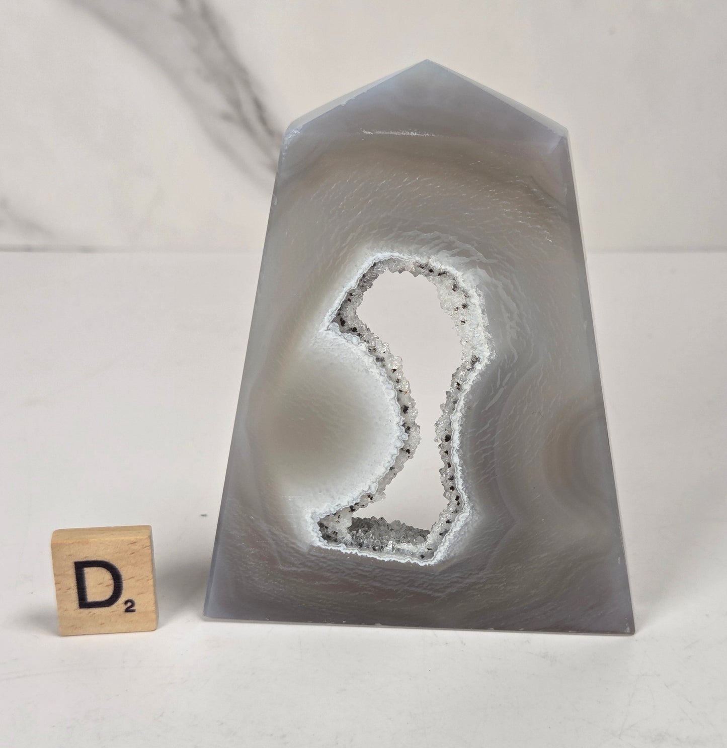 A polished natural agate crystal point with a prominent dendritic cavity, displayed on a white surface next to a small Scrabble tile showing the letter 'D'.