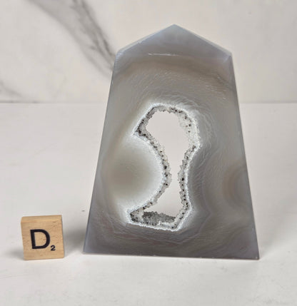 A polished natural agate crystal point with a prominent dendritic cavity, displayed on a white surface next to a small Scrabble tile showing the letter 'D'.