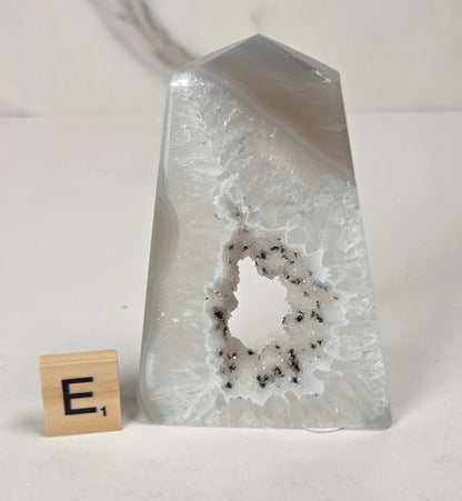 A clear, pointed crystal or glass geode-like object with a white frosted interior and dark speckles around the cavity, shown on a white surface with a small wooden letter tile beside it.