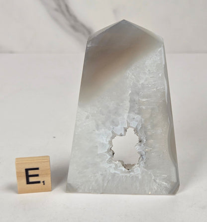 A clear, translucent pointed crystal or stone object with a rough, hollowed-out circular opening near the lower center, standing on a light surface next to a small wooden block with the letter E.