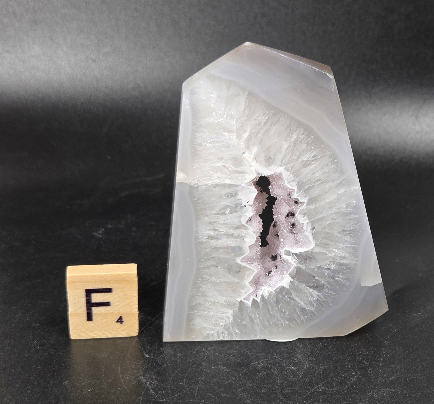A polished agate crystal point with a natural geode cavity, shown next to a small wooden Scrabble tile on a dark gray background.