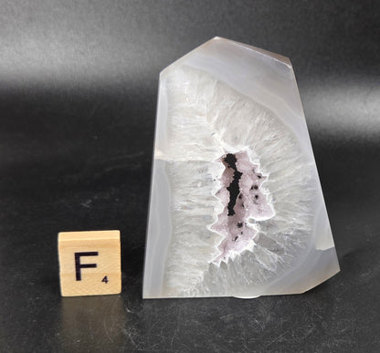 A polished agate crystal point with a natural geode cavity, shown next to a small wooden Scrabble tile on a dark gray background.