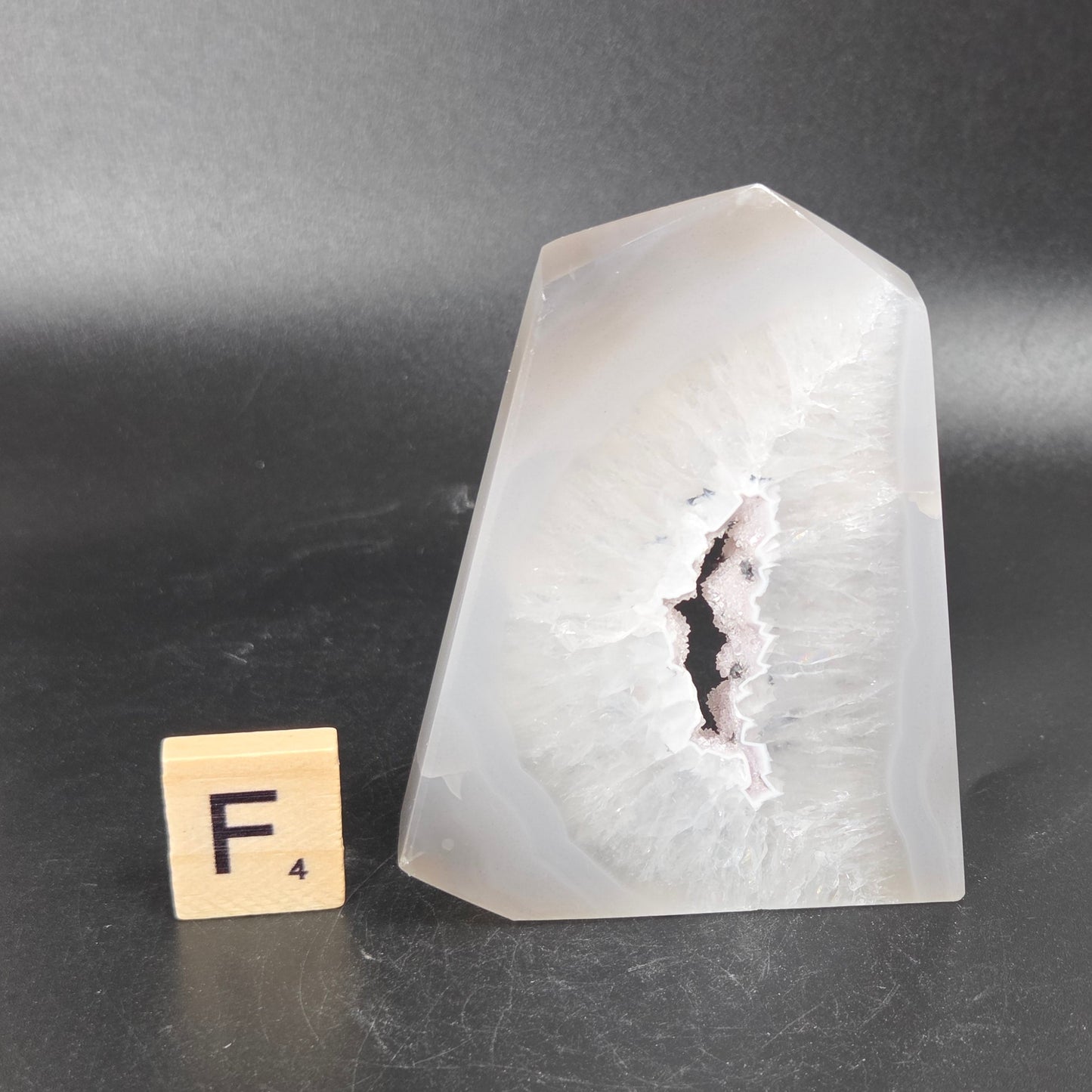 A polished white agate crystal with a natural geode-like cavity, shown on a dark gray background next to a small wooden letter tile with the letter F.