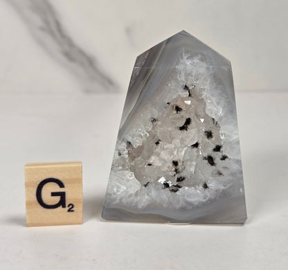 A clear quartz crystal geode point with white and black inclusions, displayed next to a wooden Scrabble tile on a light marble surface.