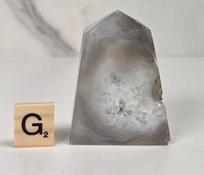 A polished agate crystal cluster stand with a pointed top, shown next to a wooden Scrabble tile marked G2 on a light marble-patterned surface.