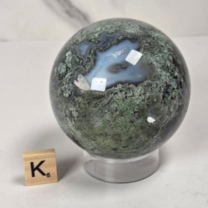A polished agate crystal sphere with green and gray banding and a light blue center, displayed on a clear acrylic stand next to a wooden Scrabble tile on a light gray surface.