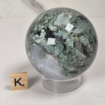 A polished geode sphere with green and gray crystalline patterns, displayed on a clear acrylic stand next to a wooden Scrabble tile on a light marble surface.
