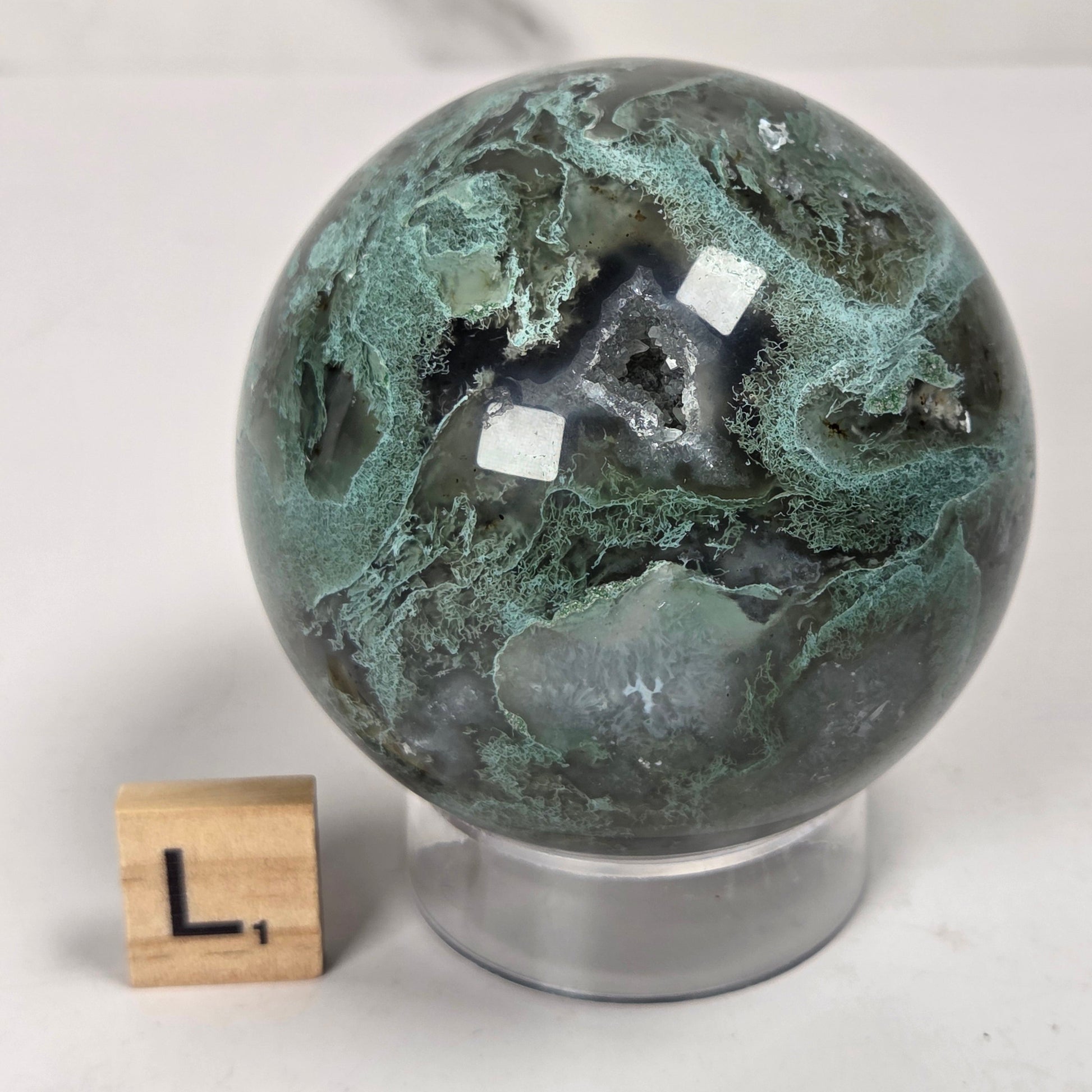 A polished green and black marbled stone sphere on a clear acrylic stand, with a small wooden block letter 'L' beside it for scale, photographed on a light surface against a softly blurred neutral background.