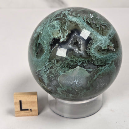 A polished green and black marbled stone sphere on a clear acrylic stand, with a small wooden block letter 'L' beside it for scale, photographed on a light surface against a softly blurred neutral background.