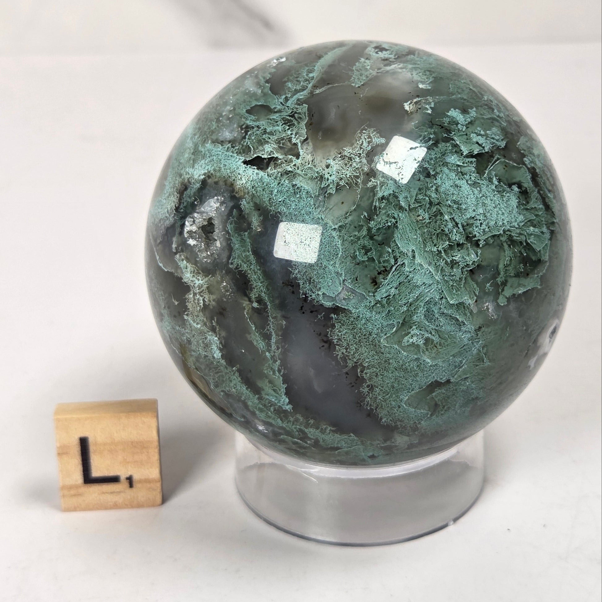 A polished green and black agate crystal sphere on a clear acrylic stand, shown next to a small wooden letter block with the letter 'L' on a light gray marble-patterned background.