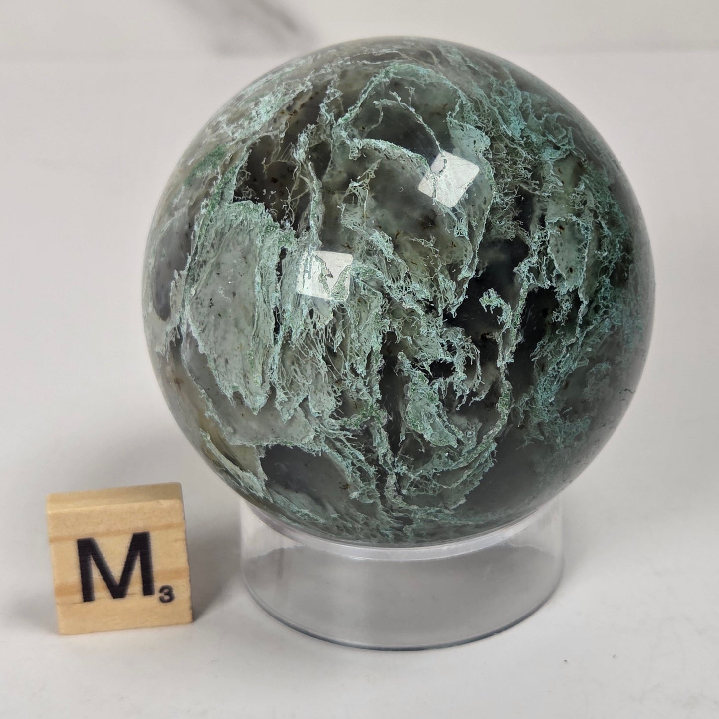 A polished green and black marbled stone sphere on a clear acrylic stand, shown next to a small wooden letter tile on a light gray marble surface.