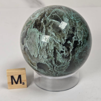 A polished green and black marbled stone sphere on a clear acrylic stand, shown next to a small wooden letter tile on a light gray marble surface.
