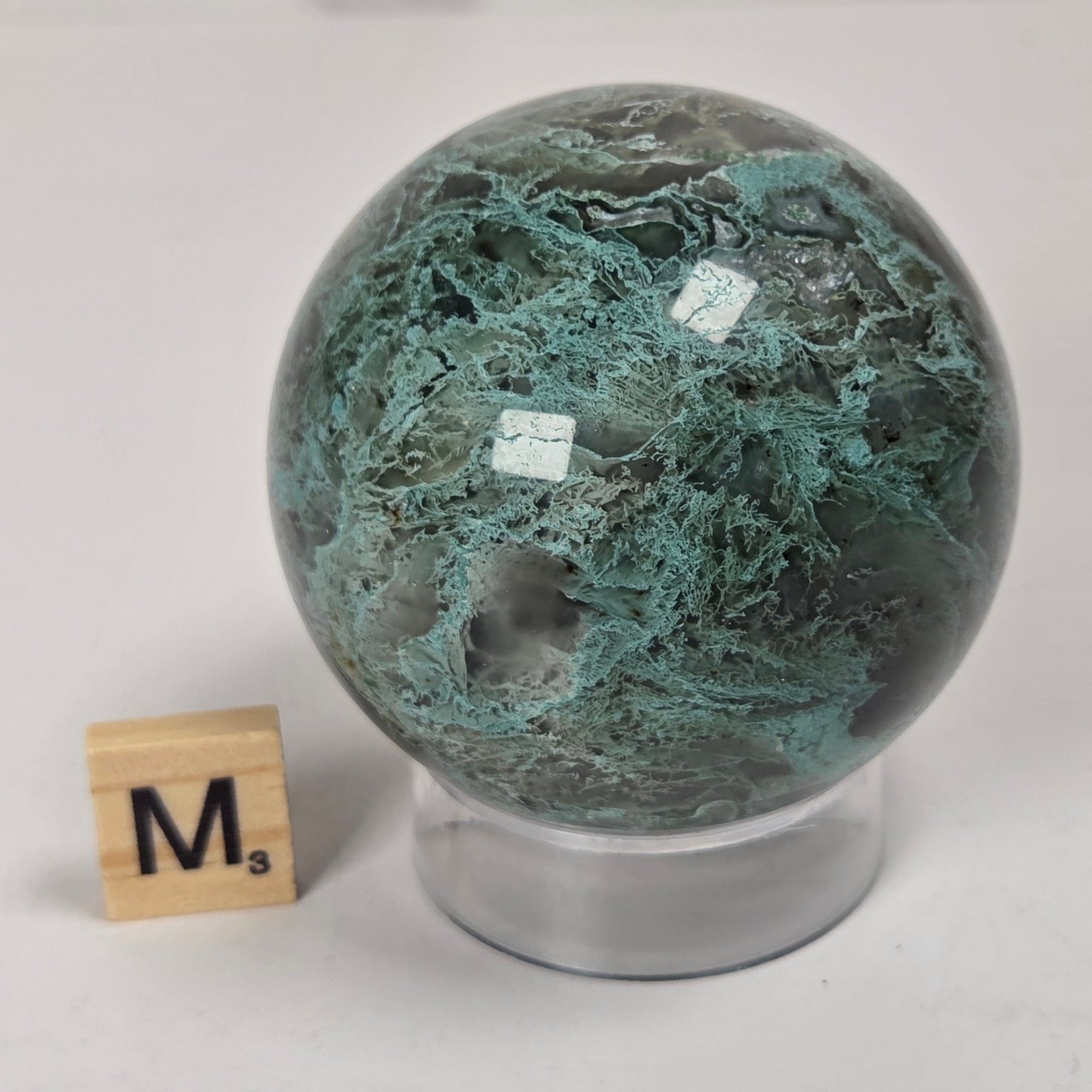 A polished green stone sphere with teal and dark marbling, displayed on a clear acrylic stand next to a small wooden Scrabble tile on a light surface.