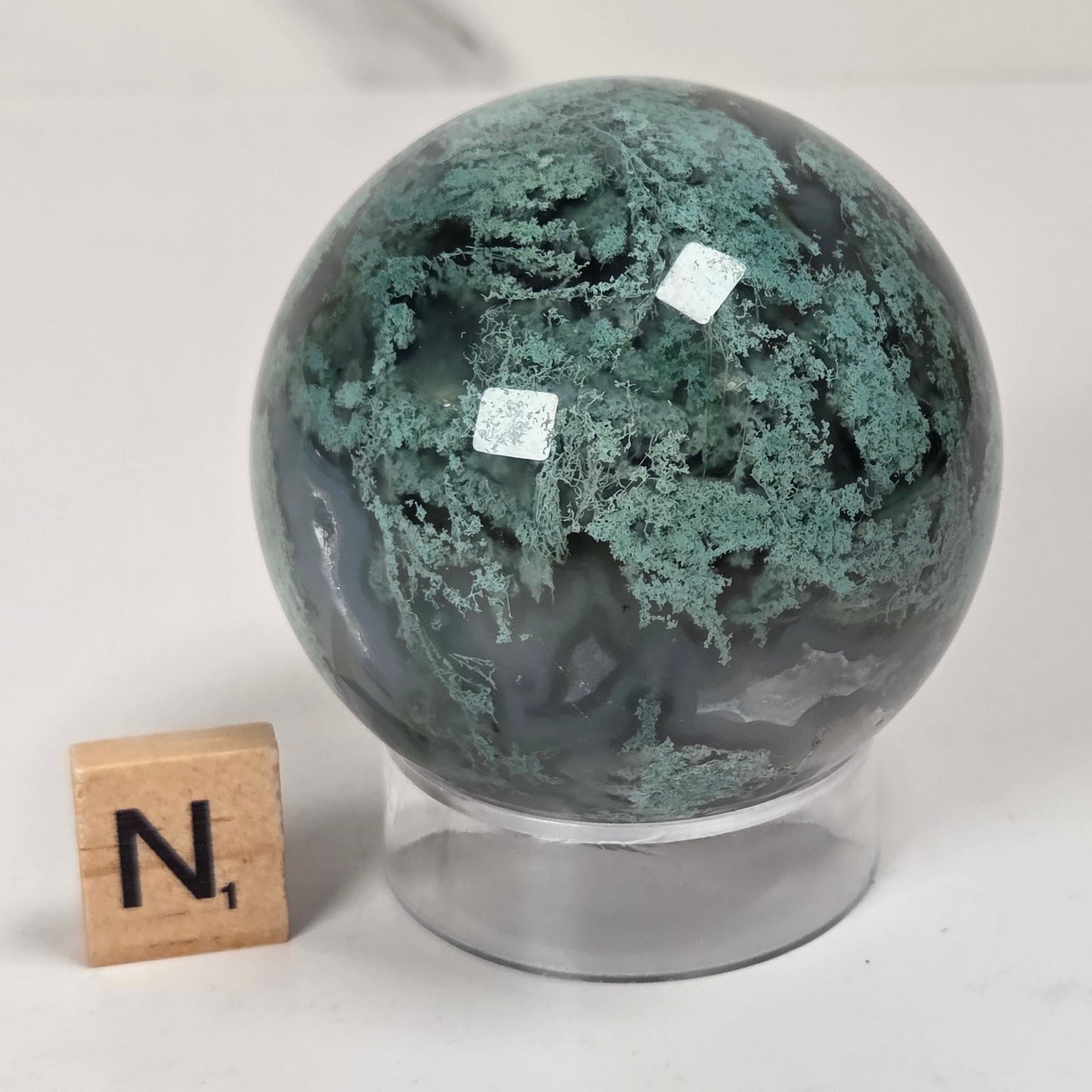A polished green and gray agate stone sphere on a clear acrylic stand, shown next to a small wooden letter tile with the letter 'N' on a white surface.