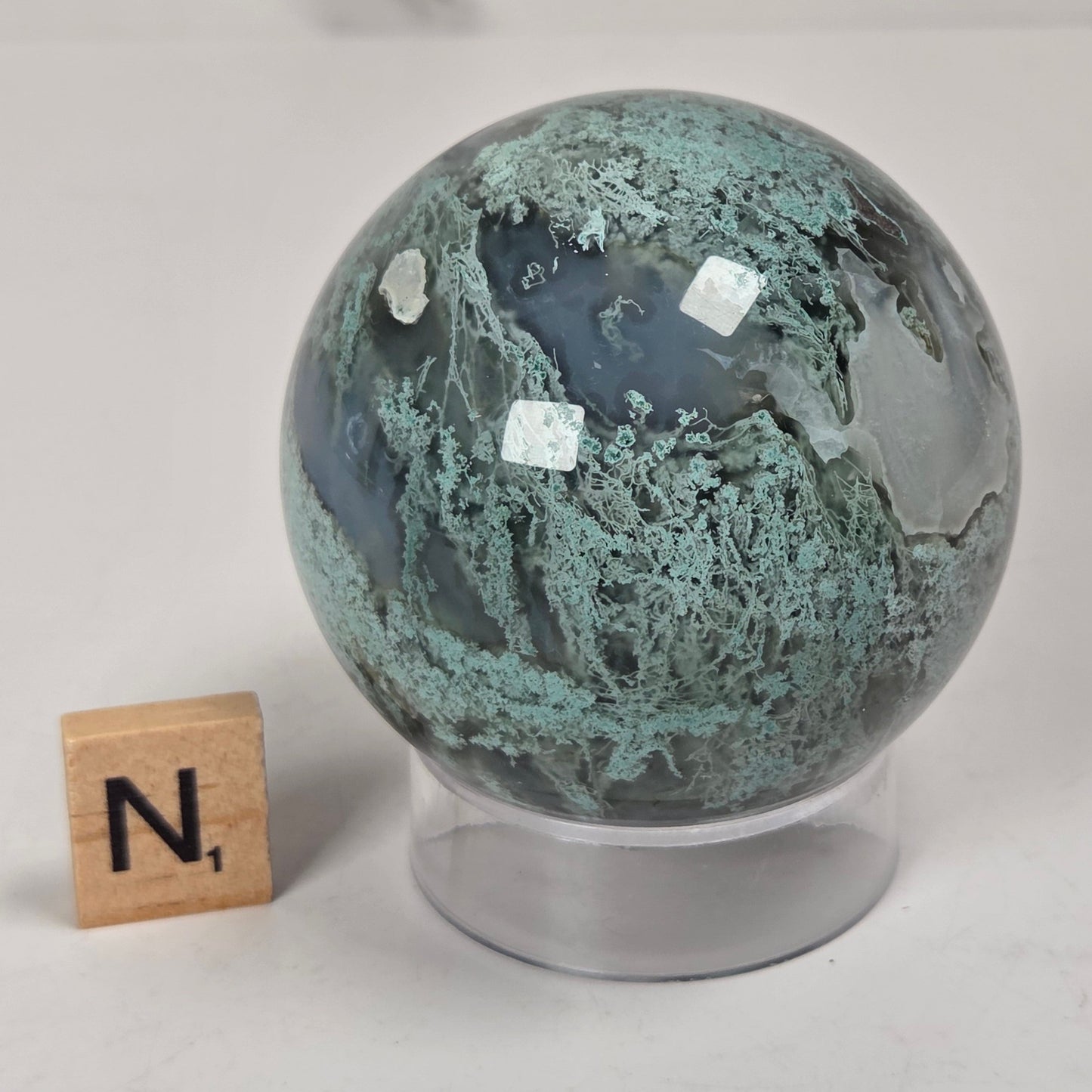 A polished round agate crystal sphere with green and gray marbled patterns, displayed on a clear acrylic stand next to a wooden letter block with the letter 'N' on a light gray surface.