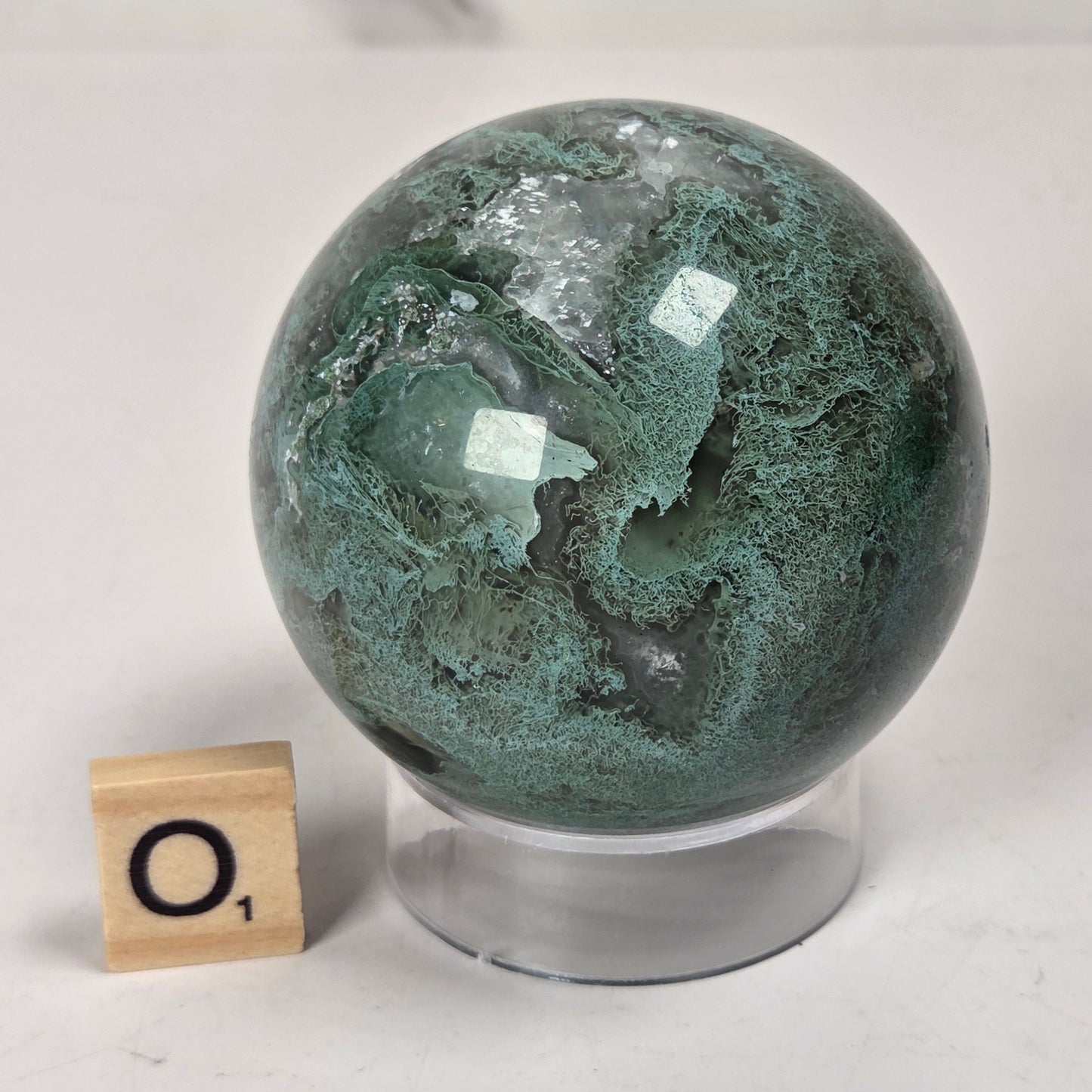 A polished green stone sphere with natural marbled veining, displayed on a clear acrylic stand next to a small wooden letter tile on a white surface.