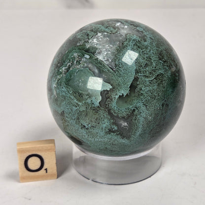 A polished green stone sphere with natural marbled veining, displayed on a clear acrylic stand next to a small wooden letter tile on a white surface.