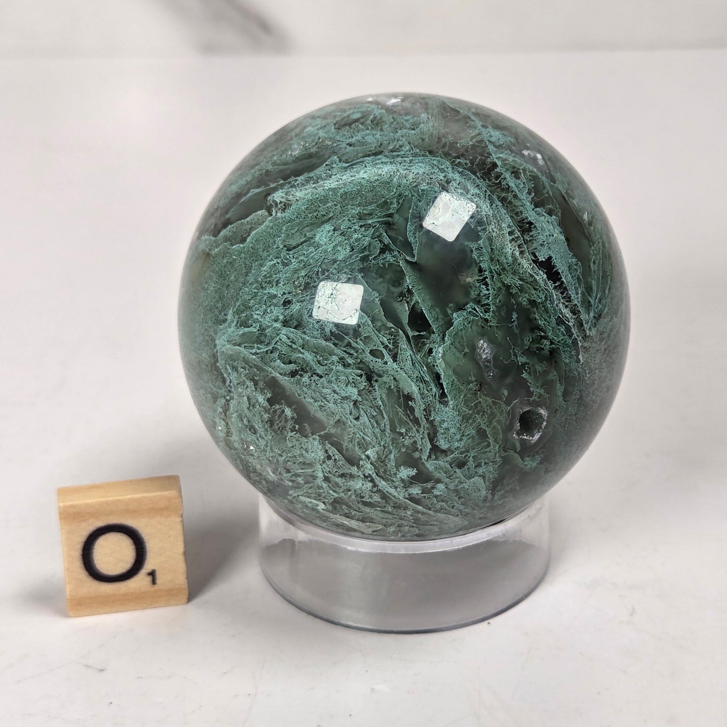 A polished green stone sphere with natural marbled veining, displayed on a clear acrylic stand next to a small wooden Scrabble tile showing the letter 'O'.