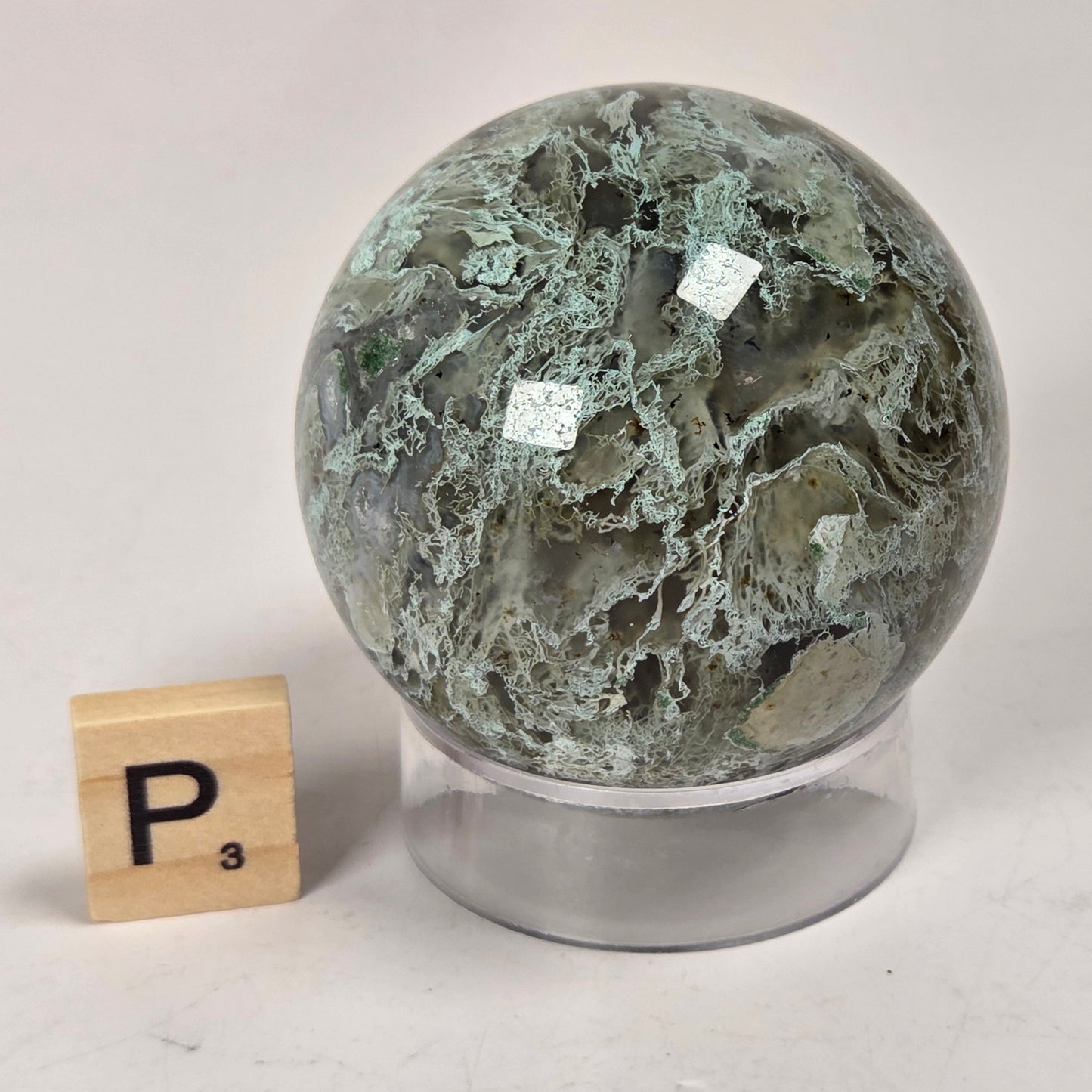 A polished green stone sphere with natural marbled veining sits on a clear acrylic display stand, photographed on a light tabletop. A small wooden Scrabble tile with the letter P is placed beside it for scale.