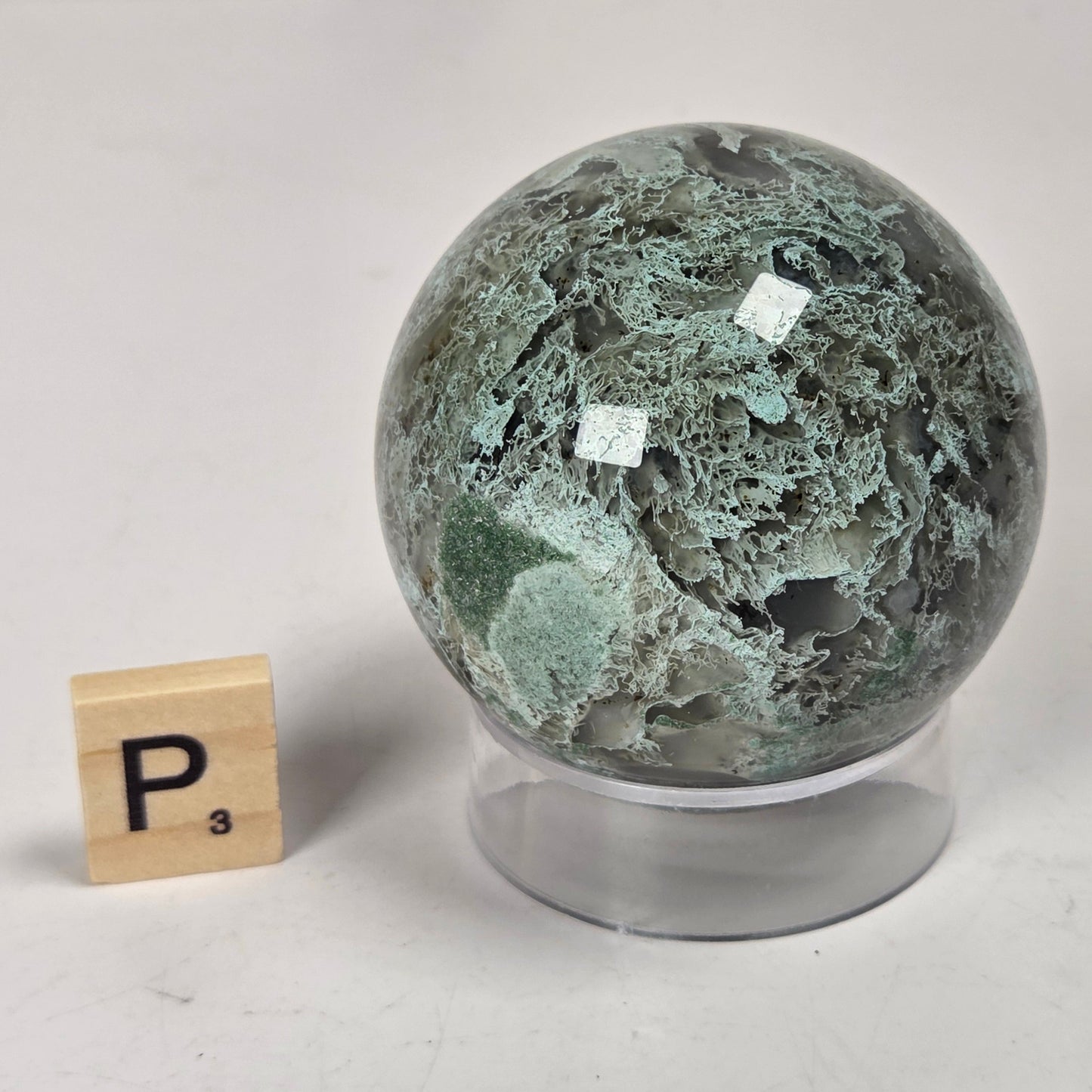 A polished green and gray stone sphere displayed on a clear acrylic stand, with a small wooden Scrabble tile showing the letter P placed to the left for size reference, on a light gray surface.