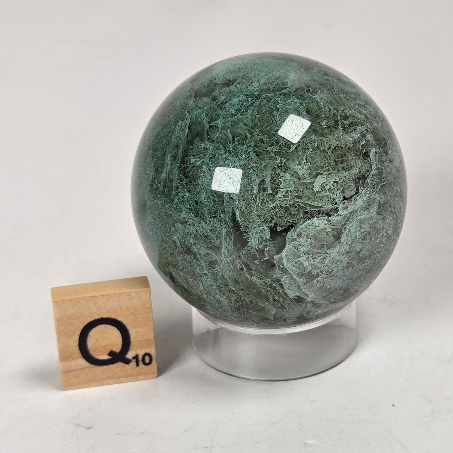 A polished green stone sphere with natural marbled veining, displayed on a clear acrylic stand next to a wooden Scrabble tile marked Q10, on a light gray surface.