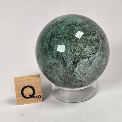 A polished green stone sphere with natural marbled veining, displayed on a clear acrylic stand next to a wooden Scrabble tile marked Q10, on a light gray surface.