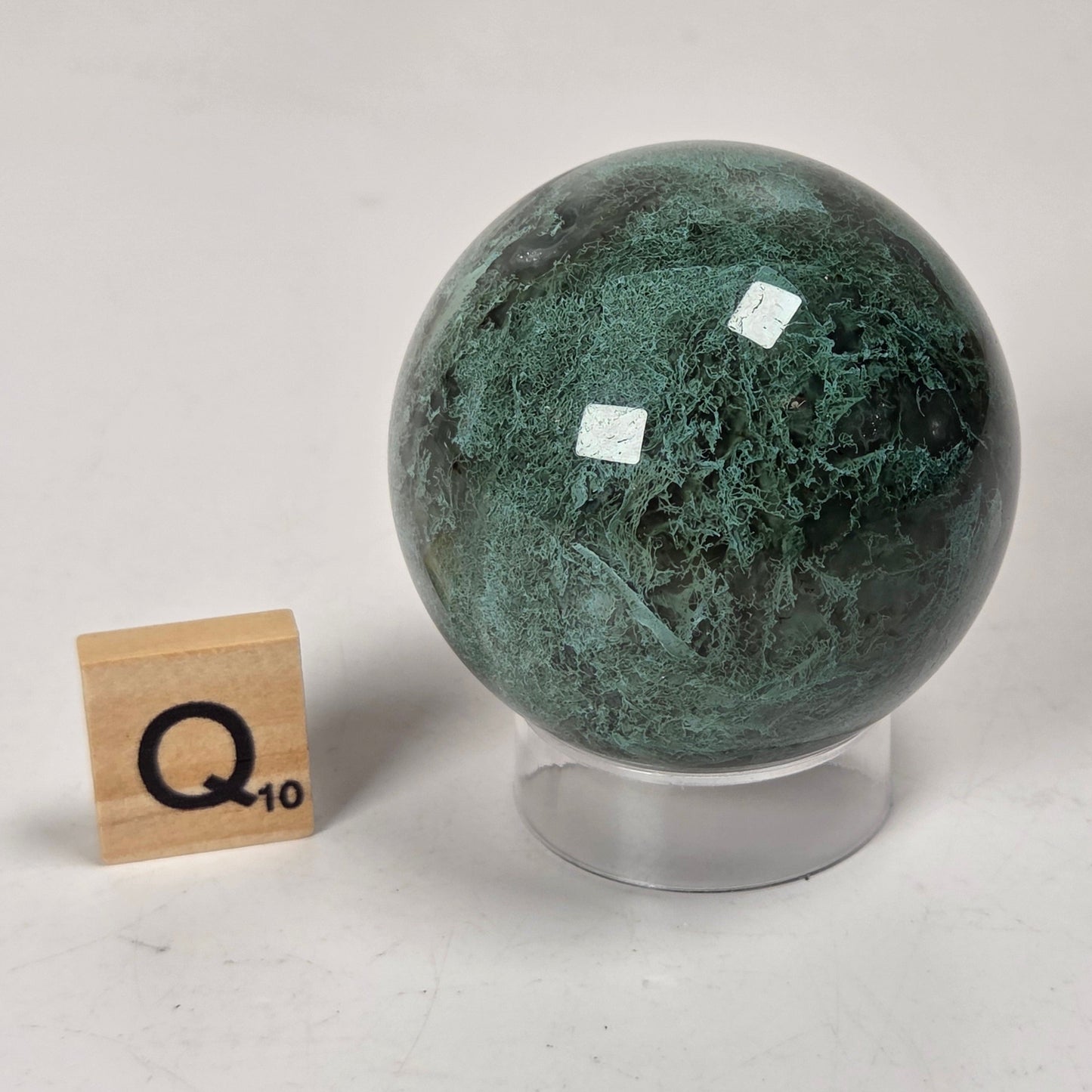 A polished green stone sphere with natural marbling sits on a clear acrylic stand on a white surface, next to a small wooden block displaying 'Q10'.