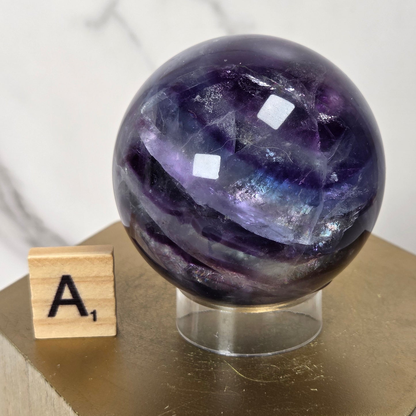 A polished purple fluorite crystal sphere on a clear acrylic stand, displayed next to a wooden Scrabble tile showing 'A1' on a gold-colored block.