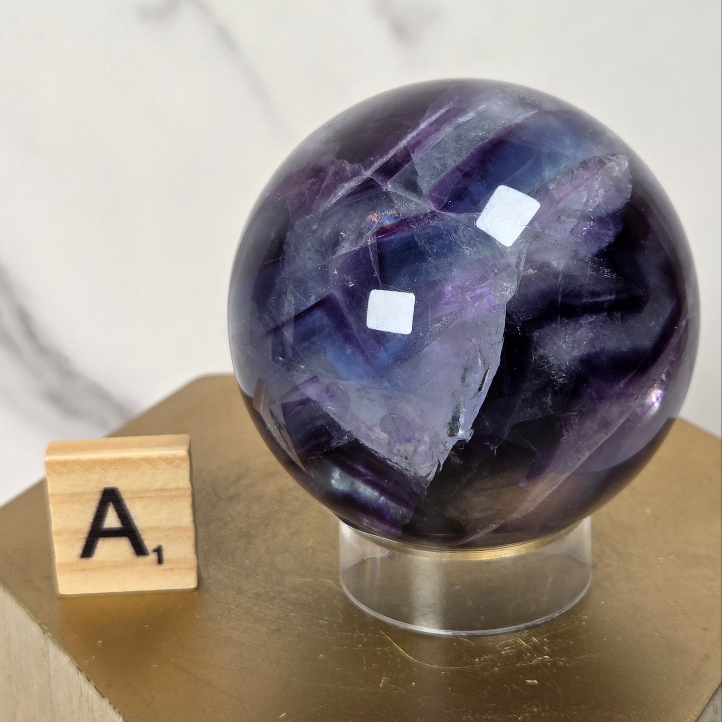 A polished purple and blue fluorite crystal sphere on a clear acrylic stand, displayed next to a wooden Scrabble tile on a gold-colored geometric pedestal.