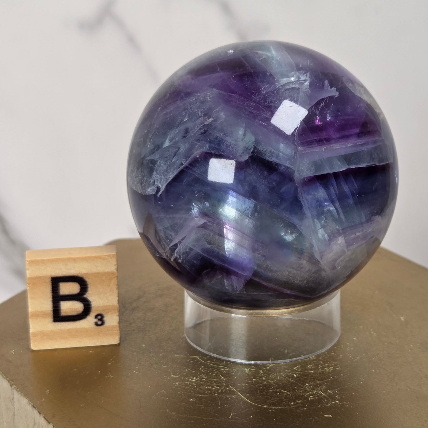 A polished purple and blue agate crystal sphere on a clear acrylic stand, displayed next to a wooden Scrabble tile on a gold hexagonal pedestal.