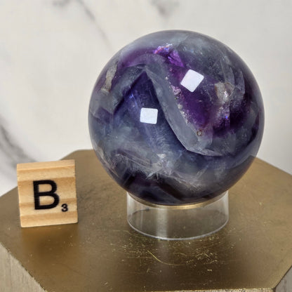 A polished purple and gray agate crystal sphere on a clear acrylic stand, displayed on a gold hexagonal pedestal next to a wooden Scrabble tile showing the letter B with a subscript 3, against a white marble background.