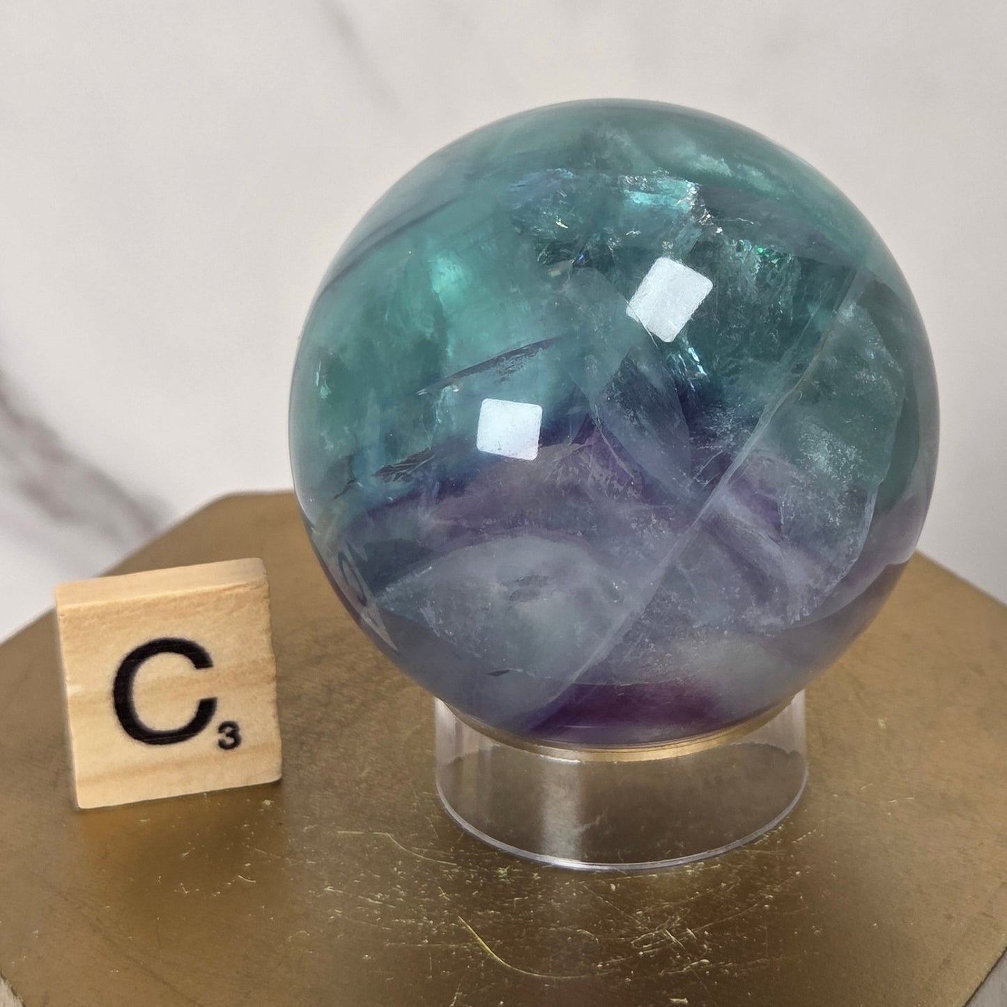 A polished round crystal sphere with swirling teal, blue, and purple tones, displayed on a clear stand atop a gold hexagonal pedestal next to a wooden Scrabble tile.