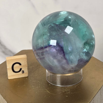 A polished round crystal sphere with swirling teal, blue, and purple tones, displayed on a clear stand atop a gold hexagonal pedestal next to a wooden Scrabble tile.